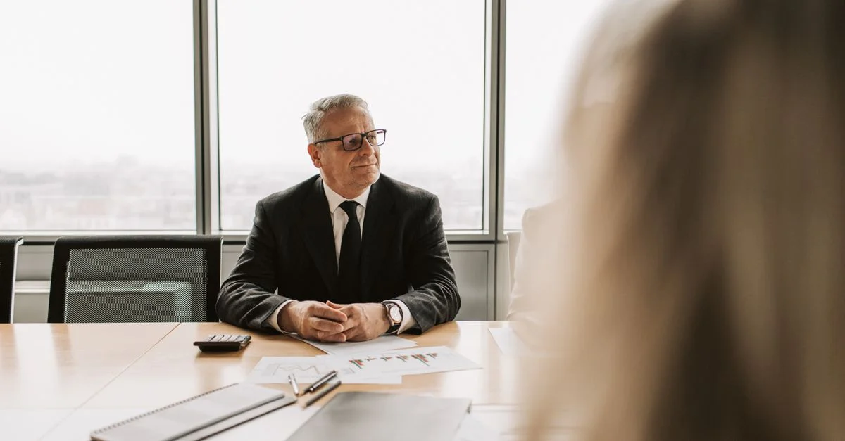 Homme d'affaires senior lors d'une réunion d'entreprise, portant un costume noir et des lunettes, à l'intérieur.