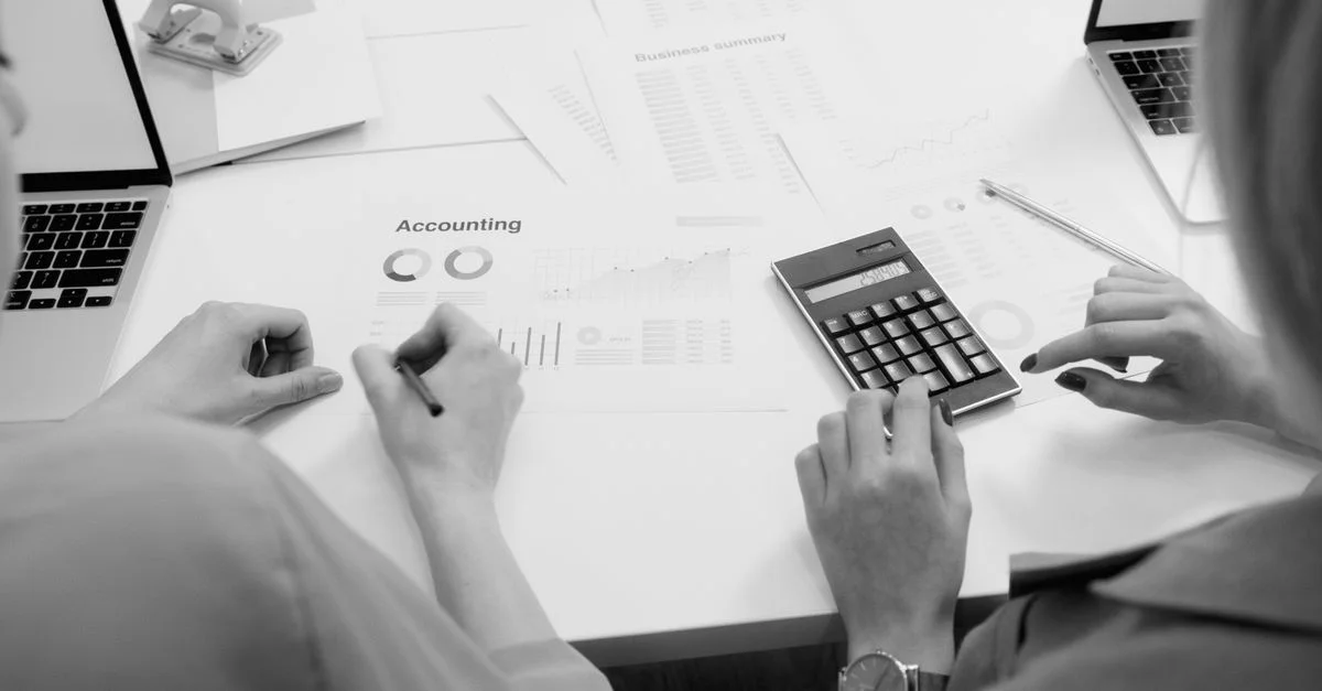 Scène de bureau en noir et blanc avec des documents comptables, une calculatrice et des ordinateurs portables.