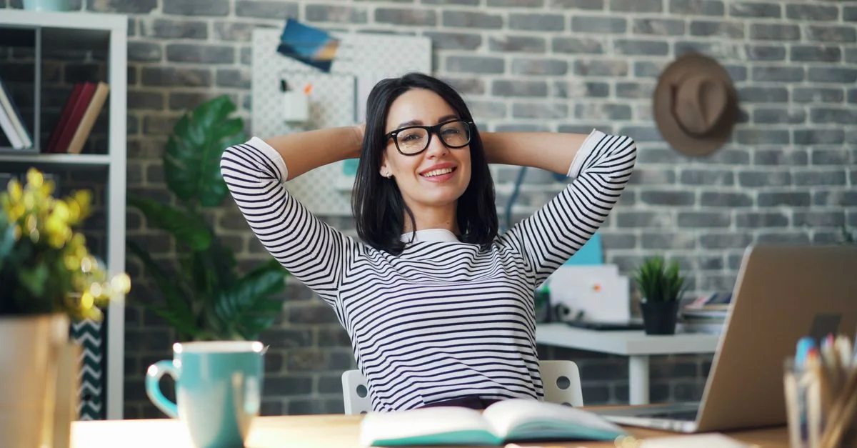 Femme souriante portant des lunettes se relaxant les mains derrière la tête à un bureau dans un bureau moderne.