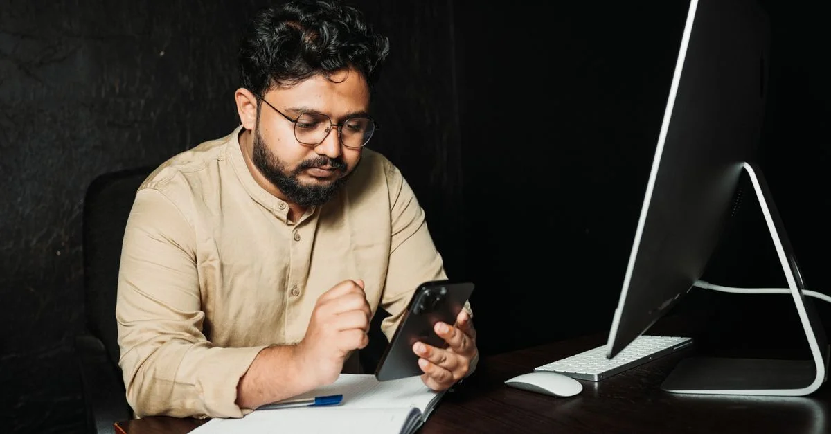 Jeune homme d'affaires vérifiant son smartphone à son bureau dans un bureau moderne, travaillant sur un ordinateur.