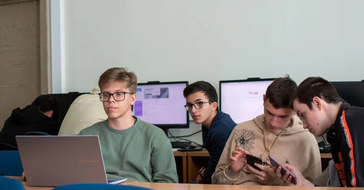Cinq adolescents engagés avec des ordinateurs portables et des smartphones dans une salle de classe moderne.