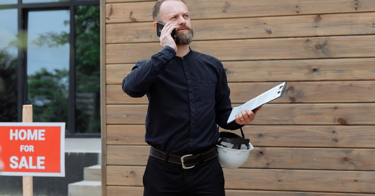 Agent immobilier passant un appel téléphonique tout en tenant des documents près d'un panneau "maison à vendre".