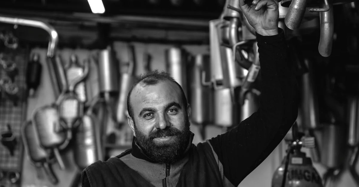 Un mécanicien souriant tenant un tuyau d'échappement dans un garage de réparation automobile. Photo en noir et blanc.