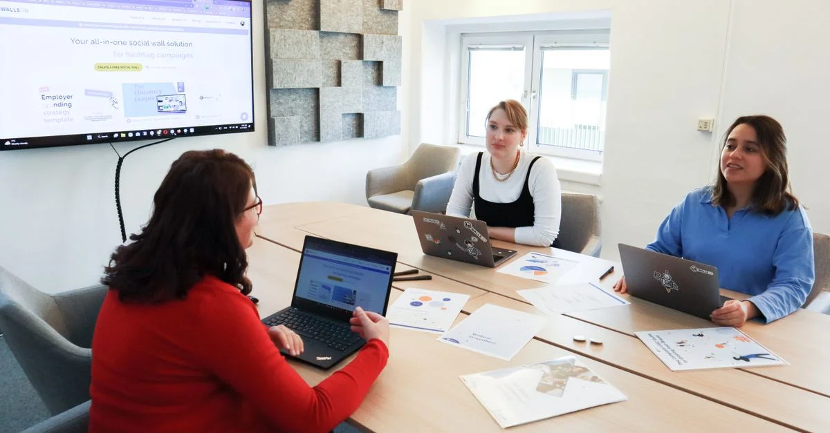 Trois femmes collaborant autour d'une table de conférence dans un bureau moderne avec des ordinateurs portables et un écran de présentation.
