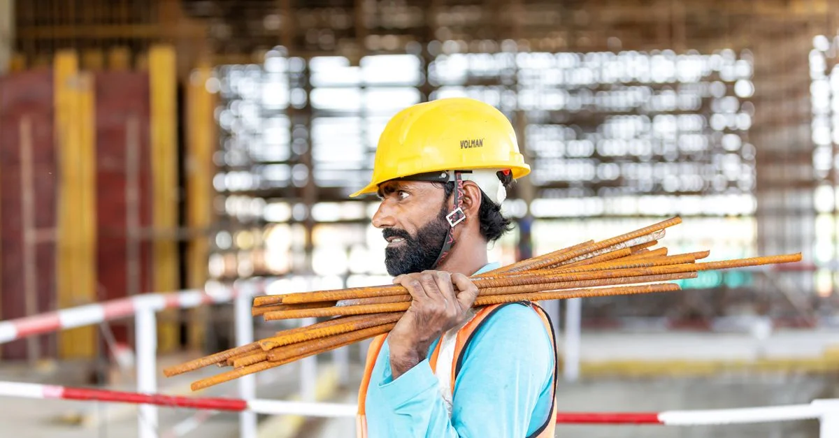 Ouvrier du bâtiment sur un chantier transportant des barres d'armature, portant un casque et un gilet de sécurité.