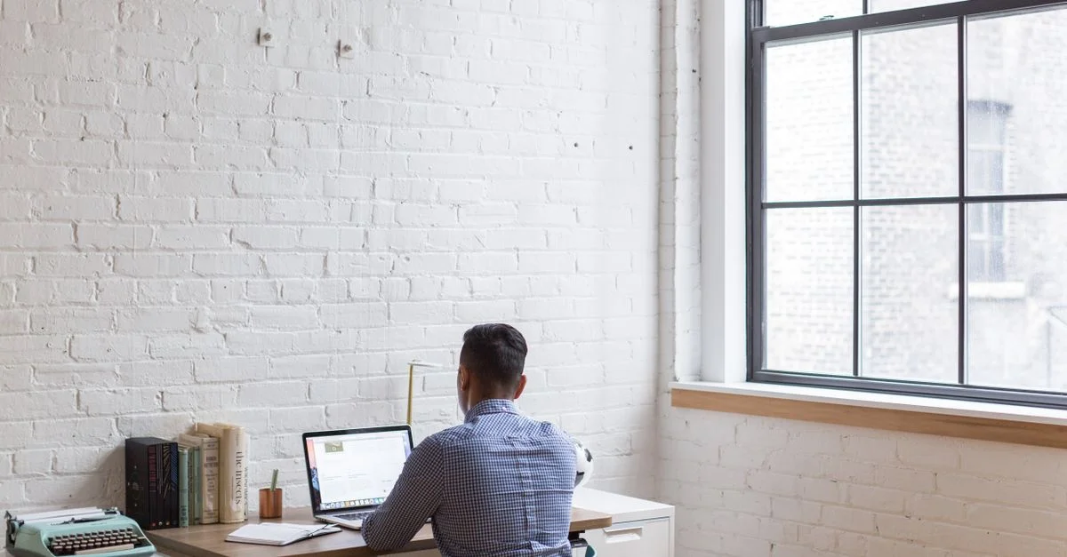 Homme adulte travaillant à distance sur un ordinateur portable dans un bureau à domicile bien éclairé avec une grande fenêtre.