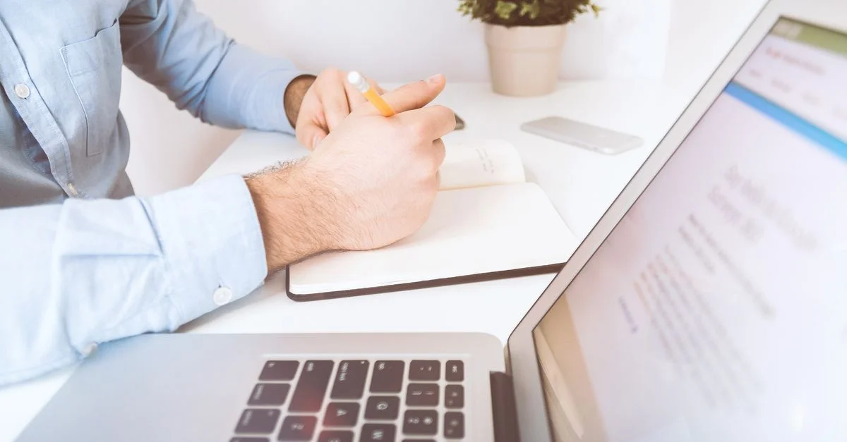 Un homme écrivant dans un carnet sur un bureau avec un ordinateur portable et un smartphone, dans un cadre de bureau moderne.
