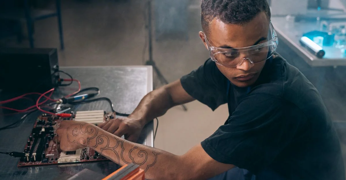 Jeune technicien avec des tatouages et des lunettes de protection travaillant sur de l'électronique dans un cadre d'atelier.