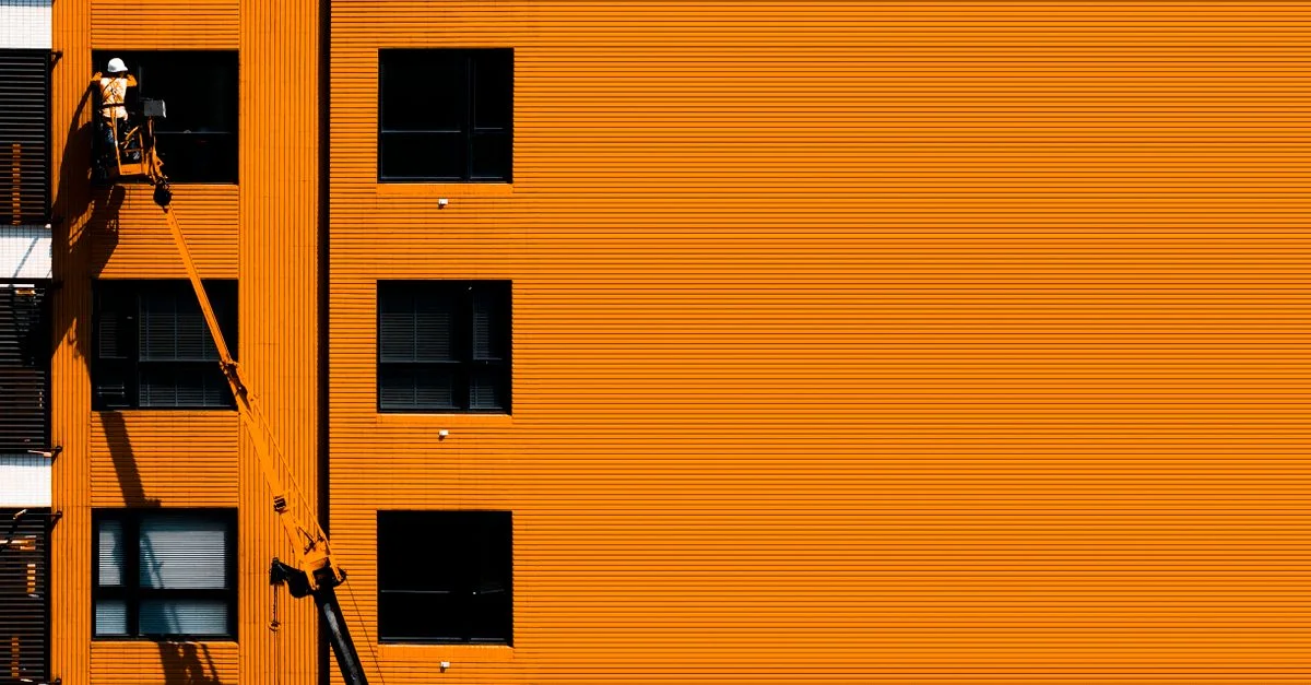 Un ouvrier du bâtiment utilisant une nacelle pour travailler sur une façade de bâtiment orange.
