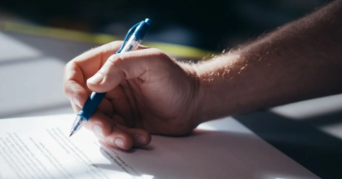 Gros plan d'une main tenant un stylo bleu pendant la signature d'un document. Idéal pour des thèmes juridiques et commerciaux.