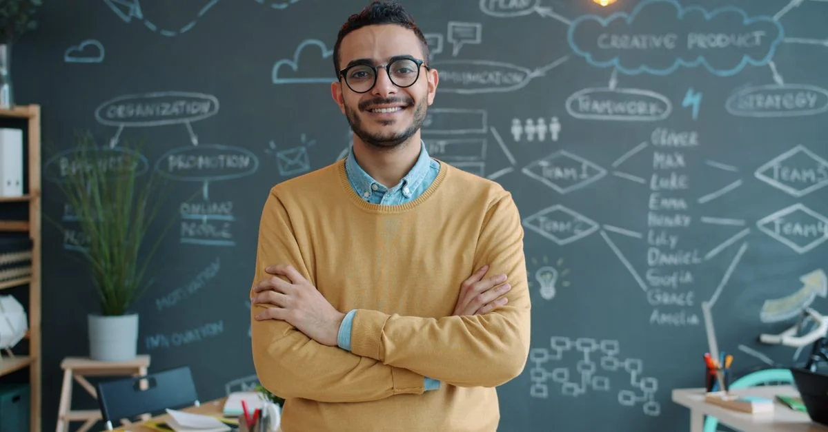 Un jeune homme souriant, vêtu de façon décontractée, se tient avec assurance dans un bureau moderne devant un tableau noir.