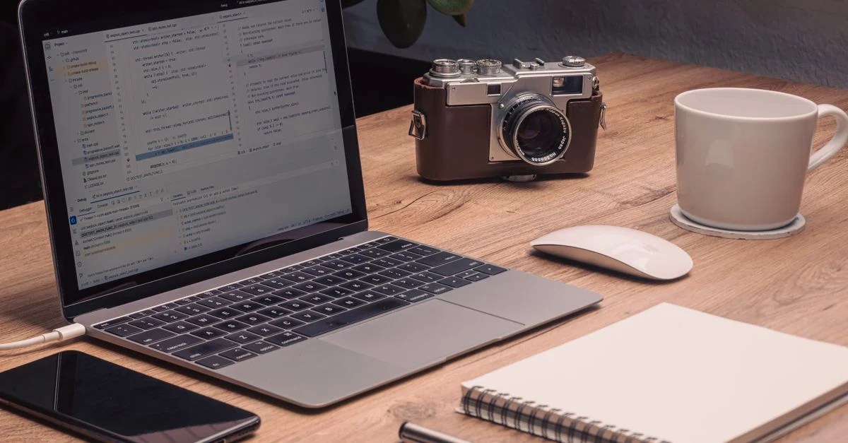Un espace de travail artistique et chaleureux avec un ordinateur portable affichant du code, un appareil photo vintage et un carnet sur un bureau en bois.