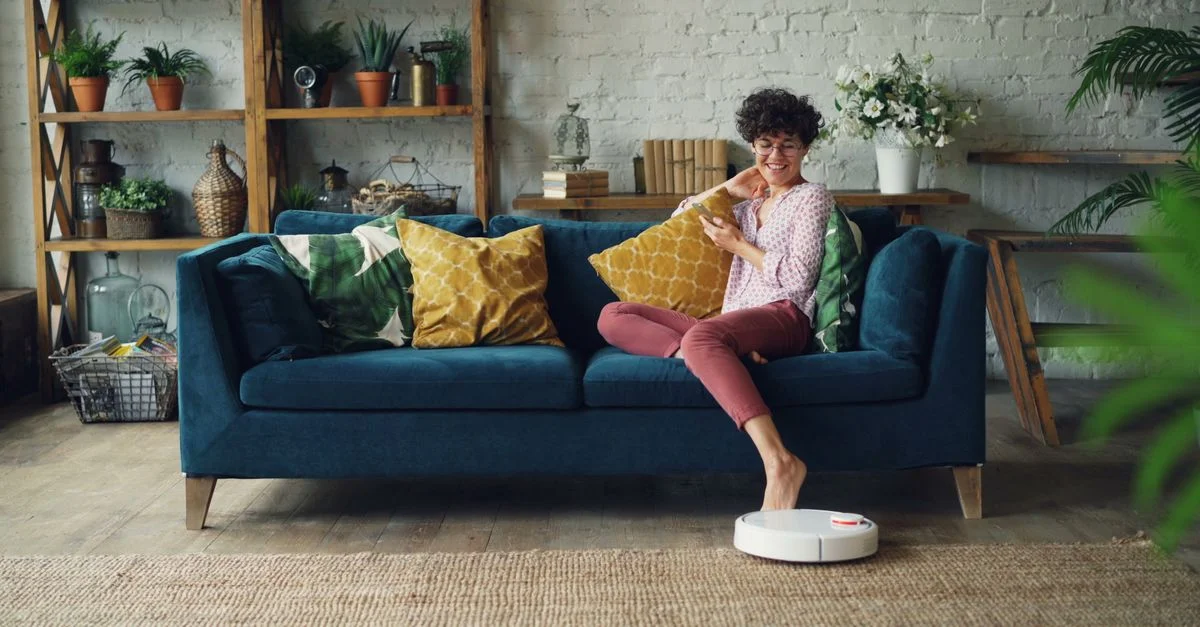 Femme se relaxant sur le canapé pendant qu'un aspirateur robot nettoie la pièce, mettant en valeur un intérieur moderne et cozy.