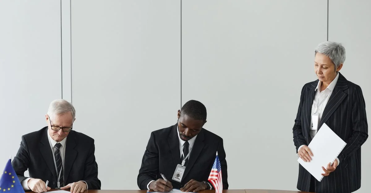 Des dirigeants signant un accord international avec les drapeaux de l'UE et des États-Unis affichés sur une table en bois.